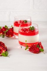 Creamy vanilla panna cotta with red jelly in beautiful glasses, fresh ripe pomegranate on white wooden background.