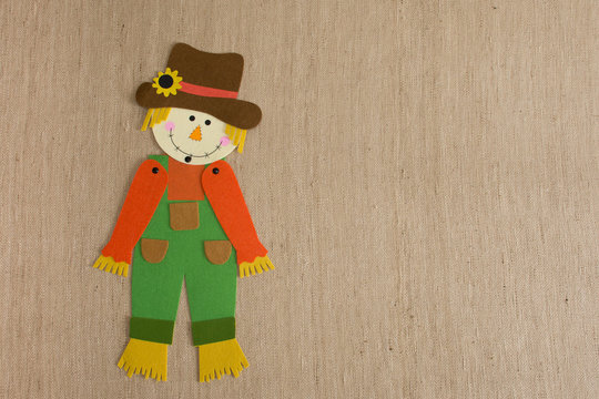 Scarecrow with Tilted Head wearing green overalls,  an orange shirt and a brown hat adorned with a sunflower.  Photographed against tan woven cloth.