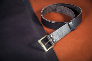 A close-up black belt made of genuine leather made by hand and a few buckles lie on the table on pieces of black and brown leather