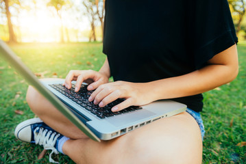 Young asian woman's legs on the green grass with open laptop. Girl's hands on keyboard. Distance learning concept. Happy hipster young asian woman working on laptop in park. Student studying outdoors.