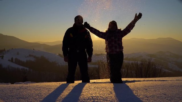Two young tourists stand in deep snow on the mountain, holding hands and cheerfully throw snow up. Man and woman. In the background, the sun sets over the mountain.
