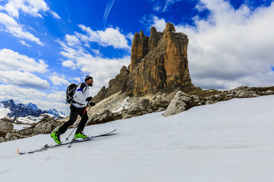 Mountaineer backcountry ski walking up along a snowy ridge with skis in the backpack. In background blue cloudy sky and shiny sun and Tre Cime, Drei Zinnen in South Tirol, Italy. 