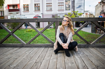 Blonde girl at fur coat sitting on wooden floor outdoor.