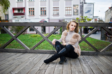 Blonde girl at fur coat sitting on wooden floor outdoor.