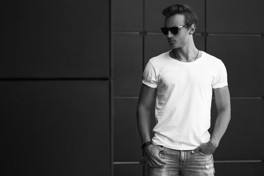 Street fashion concept. Portrait of a young and handsome man in a white t shirt and trendy glasses posing over black modern background. Close up. Street shot. copy space