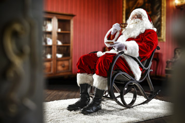 Obraz premium Santa Claus sitting in a wooden chair. Interior of the house with red walls and old furniture. Blurred wall fragments on first background.