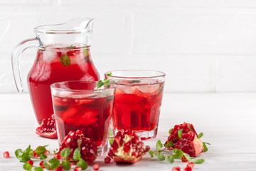Pomegranate cocktail with ice and mint in beautiful glasses and jug, fresh ripe pomegranate on white wooden background. Sweet red juice. Close up photography. Selective focus