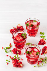 Pomegranate cocktail with ice and mint in beautiful glasses and jug, fresh ripe pomegranate on white wooden background. Sweet red juice. Close up photography. Selective focus