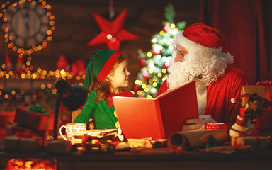 santa claus reads book to little elf by Christmas tree