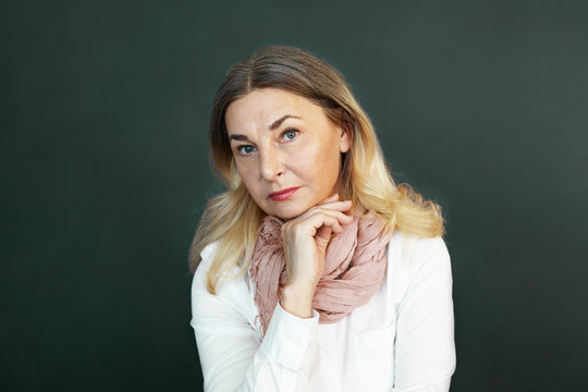 Picture of 60 year old senior female teacher wearing yellow scarf around her neck, holding hand on chin and looking at camera with serious strict expression, posing at blank copyspace studio wall