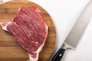 Raw beef steak on the round wooden board with kitchen chef knife on the white marble