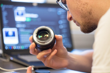 Junger Mann hält Objektiv in der Hand, Laptop im Hintergrund