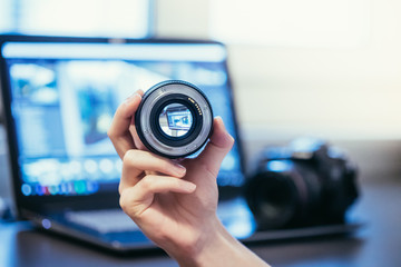 Objektiv wird in der Hand gehalten, Laptop im und Spiegelreflex Kamera im Hintergrund