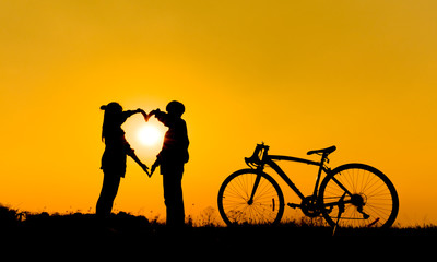 Obraz premium Two silhouettes of people making a heart shape with their hands in front of a setting sun, with a bicycle to the right.