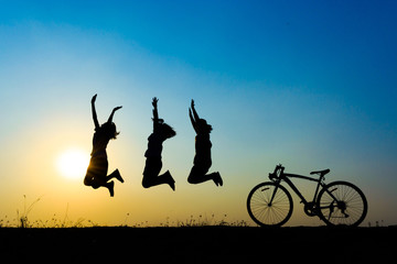 Three people are silhouetted jumping in the air in front of a sunset, with a bicycle to the right.
