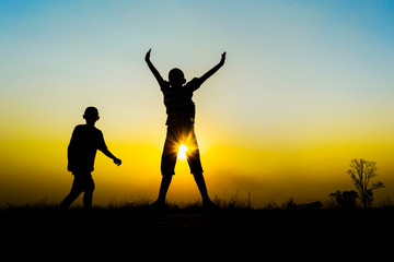  two people silhouetted against a bright orange sunset. One person is jumping with arms raised.