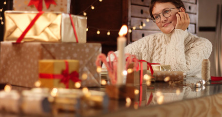 dreaming woman in thick white cable sweater wrapping presents on the background of Christmas decorations and lights