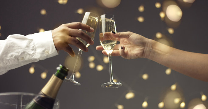 Close up of dark skinned man and caucasian woman toasting with glasses of sparkling wine on gray background with lights and lens flare