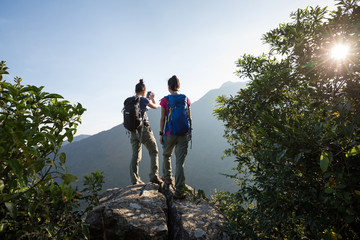 two young women backpackers enjoy the view on cliff's edge in the sunrise