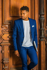 African American Businessman working in New York. Wearing blue suit, white T shirt, college student with little goatee, standing by vintage library door on campus, carrying laptop computer, thinking..