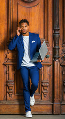 African American Businessman working in New York, wearing blue suit, unbuttoned, white undershirt, white sneakers, standing against vintage library door, working on laptop computer, talking on phone..