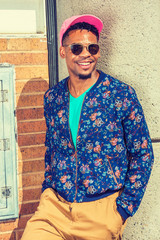 African American Man Street Fashion in New York, wearing blue patterned, collarless jacket, green V neck shirt, yellow brown pants, pink cap, sunglasses, standing against wall under sun, smiling..