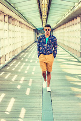 Fototapeta na wymiar African American college student, wearing blue patterned jacket, yellow brown shorts, white sneakers, sunglasses, walking on walkway with glass walls, ceiling, wooden floor on campus in New York. .
