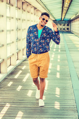 African American college student, wearing blue patterned jacket, yellow brown shorts, white sneakers, sunglasses, walking on walkway with glass walls, ceiling, wooden floor on campus in New York. .