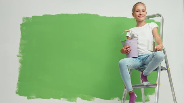 Child sitting on ladder with bucket of paint and a brush in her hand near green collor wall
