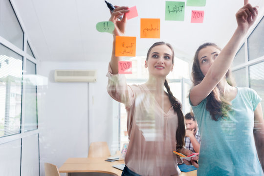 Two proficient female co-workers posting sticky notes in the interior of a shared office space