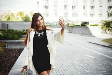 Fototapeta premium Beautiful young fashionable woman in a black short dress and white cardigan posing in the city. Fashion photo