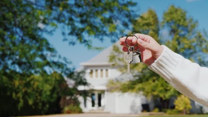 A hand with a keychain in the shape of a small house and keys. Against the background of a typical house in the American suburban style - Powered by Adobe