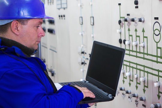 Technician in blue with laptop reading instruments in power plant control center