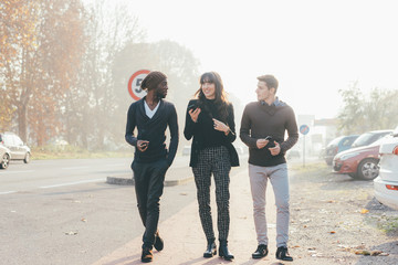 Three multiethnic woman and men friends walking outdoor in city back light, chatting and holding smart phone - friendship, social network, conversation concept