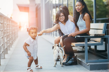 Three happy multinational best girl friend with toddler baby girl and dog sitting on the bench smiling and talking over shopwindow background. Little pulls her mother by the hand. Sun glare effect