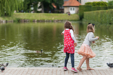 Young girls feeding birds by the city pond. Throwing bread to ducks. Pretty colorful dresses and skirts, long hair.