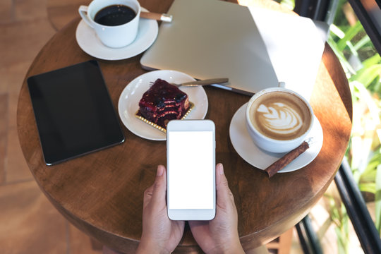 Top view mockup image of hands holding white smartphone with blank screen , tablet , laptop , coffee cup and cake on wooden table in cafe