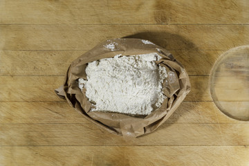 Brown paper sack of white flour in the center of rough wooden rectangular cutting board directly from above 