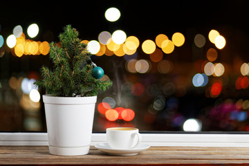 Small Christmas tree in a flowerpot decorated with one Christmas ball on a wooden window sill. Nearby is a cup of coffee. Outside the window are colored lights on blurred bacground.