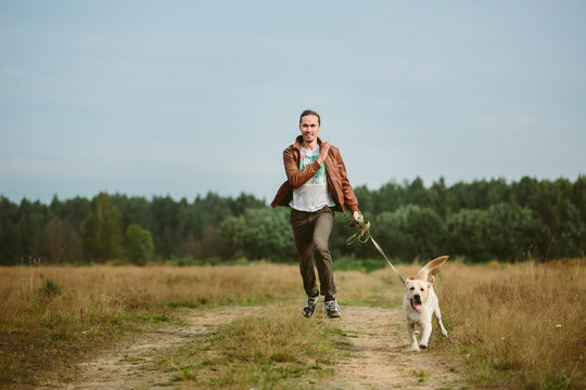 A Happy Man Running With A Dog On Country Road On A Meadow. A Man And A Dog Are Looking At Camera