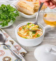 Chicken orzo soup in a white crock on wooden background. Italian soup with orzo pasta. Chef's hand holding a ladle. Bread. Glass of wine.