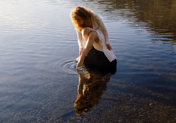 Lady with long wavy red hair crouched on a rock reflected in still lake