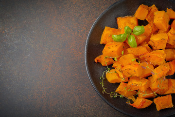 Fried pumpkin slices with herbs and pepper, olive oil in a black ceramic plate on  dark concrete or stone background. Selective focus. Top view.