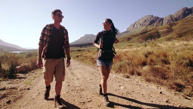 Video of young man and woman talking and walking on a dirt road. Couple with backpack hiking in nature.