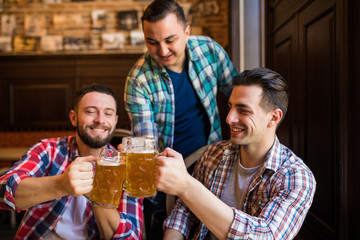 Three young men in casual clothes are smiling and clanging glasses of beer together while sitting in pub