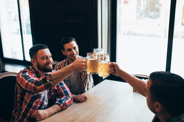 Three young happy men in casual clothes are smiling and clanging glasses of beer together while sitting in pub