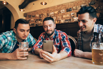 Three young men in casual clothes are talking, using a smart phone and drinking beer while sitting at bar counter in pub