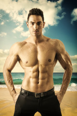 Obraz premium Attractive muscular man by the sea, shirtless looking at camera, standing in the beach