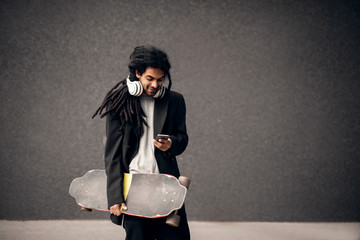 Stylish young dreadlocks hipster skater with headphones holding skateboard near the grey wall and looking on mobile in a suit. © Dusan Petkovic