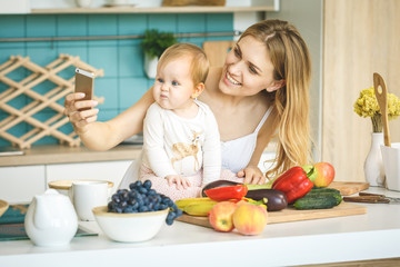Obraz na płótnie Canvas I love selfie! Young mother looking at camera and smiling, cooking and playing with her baby daughter in a modern kitchen setting. Healthy food concept.